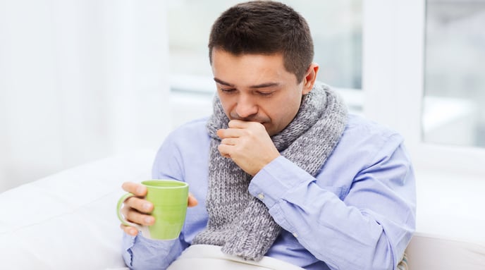 Man with a scarf around his neck coughing and holding a hot beverage
